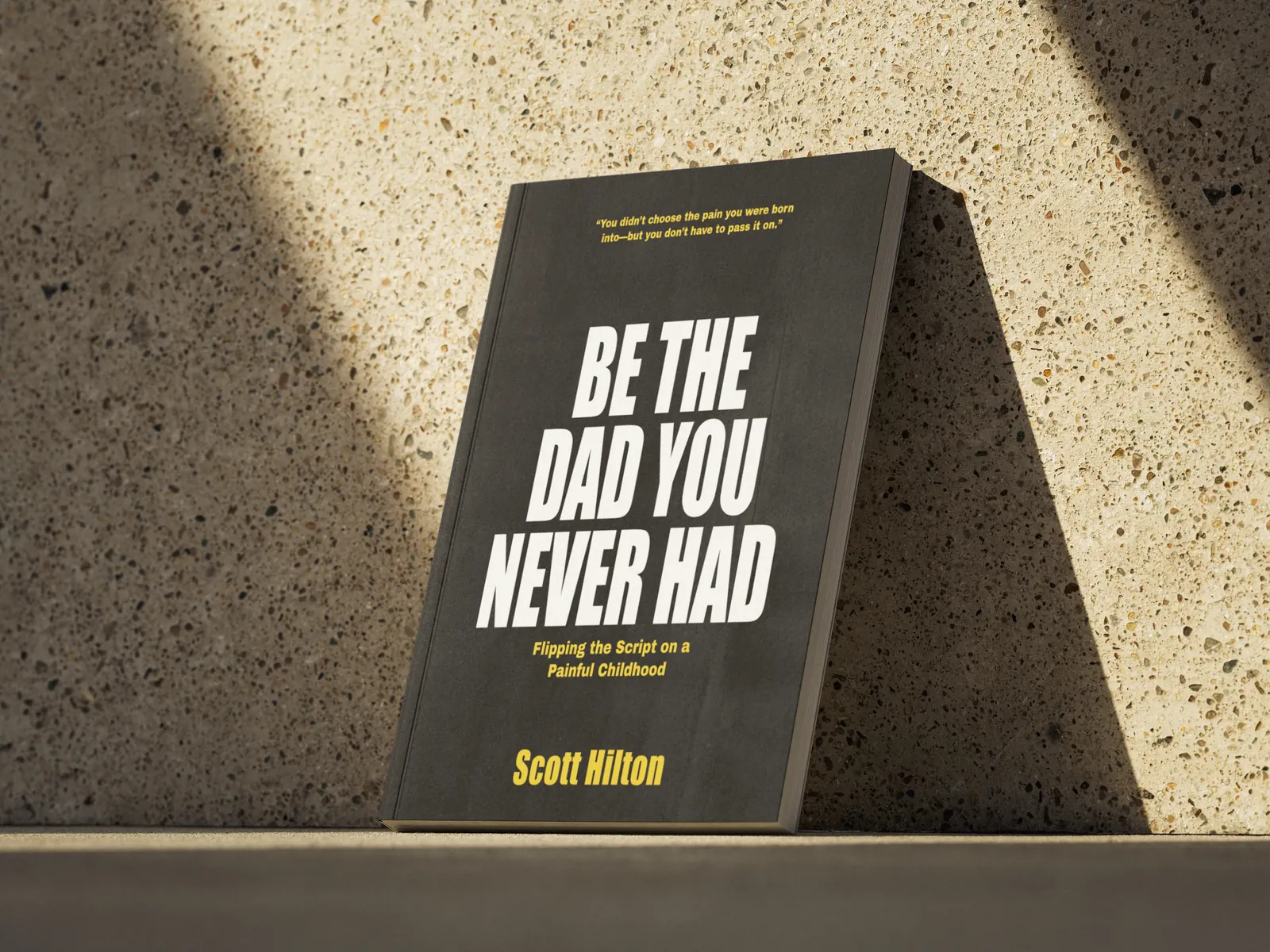 Be the Dad You Never Had by Scott Hilton, a black paperback leaning on a sunlit stone wall