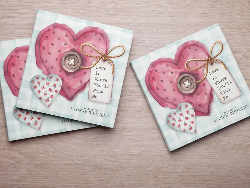 Two copies of Love Is Where You'll Find Me by Yvonne Bienvenu, gingham covers with a stitched pink heart, on a wood table